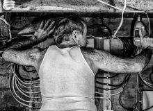 A harshly lit black and white image of a man in his basement drilling into a beam above him, the wires of the electrical panel seemingly protruding from his ribs. The texture of his clothes and his skin and hair mirror the wood, wire and steel of his environment.