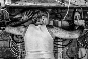 A harshly lit black and white image of a man in his basement drilling into a beam above him, the wires of the electrical panel seemingly protruding from his ribs. The texture of his clothes and his skin and hair mirror the wood, wire and steel of his environment.